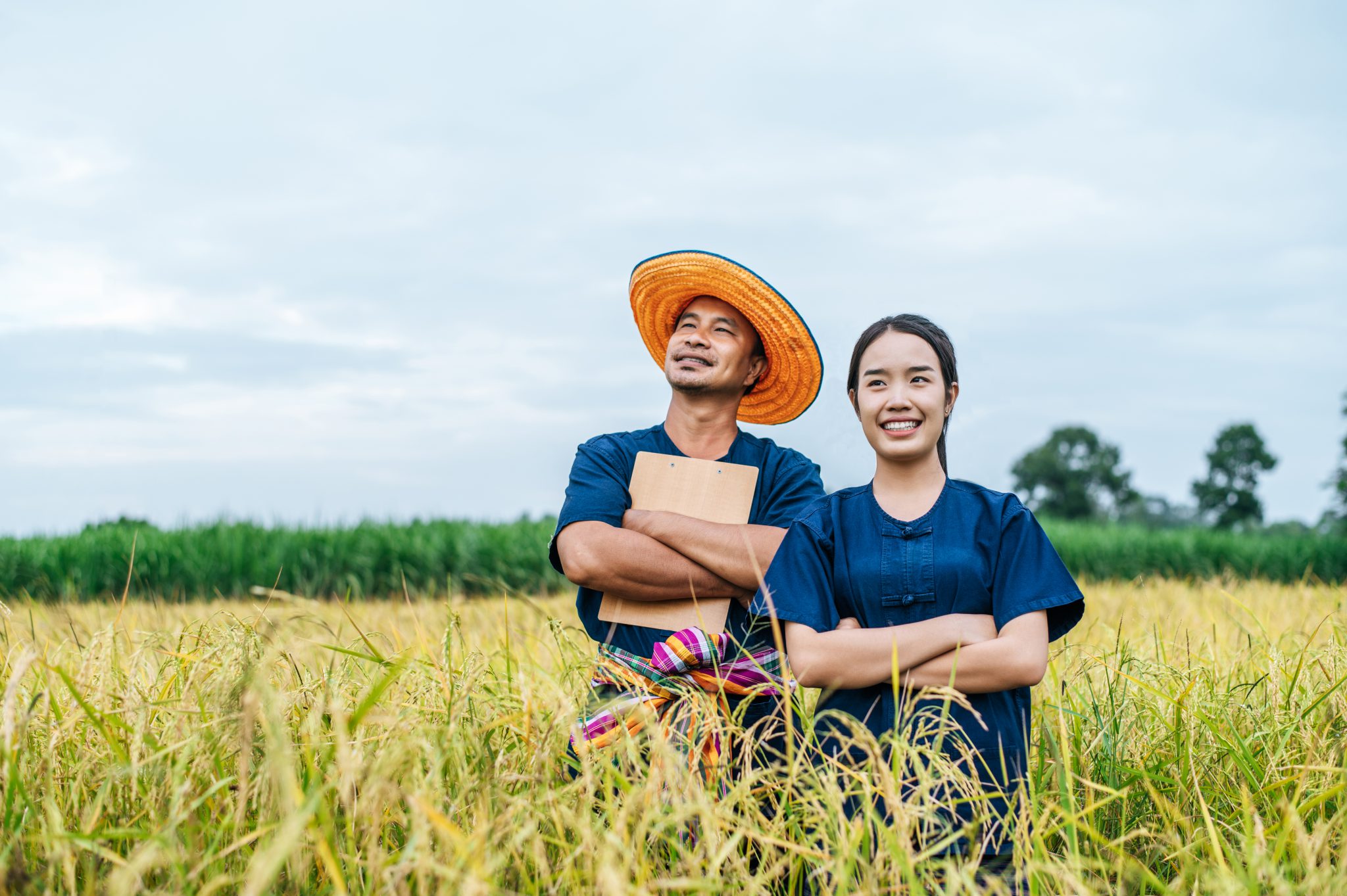Portrait Asian Middle aged man wearing straw hat and loincloth and young woman farmer stand and cross arms in rice field together,  they are smile with happiness copy space